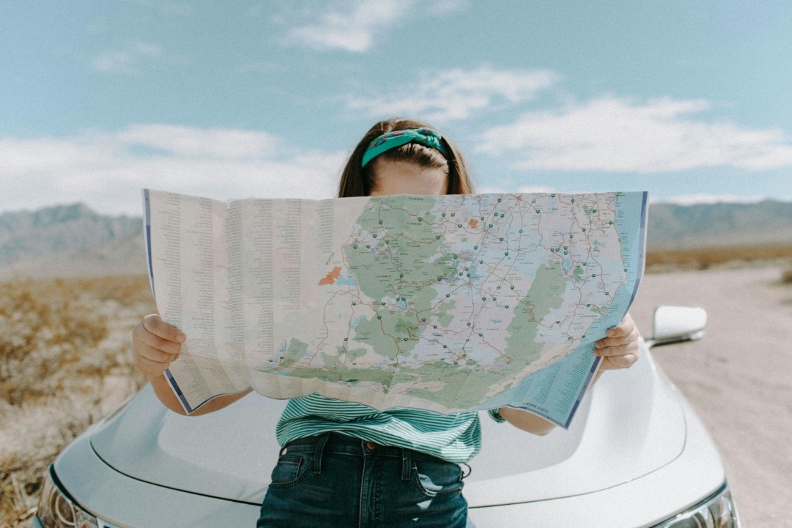 Junge Frau lehnt auf einem Auto und hält eine Landkarte in den Händen. Das Gesicht ist mit der Karte verdeckt. Im Hintergrund Bergsilhouette.