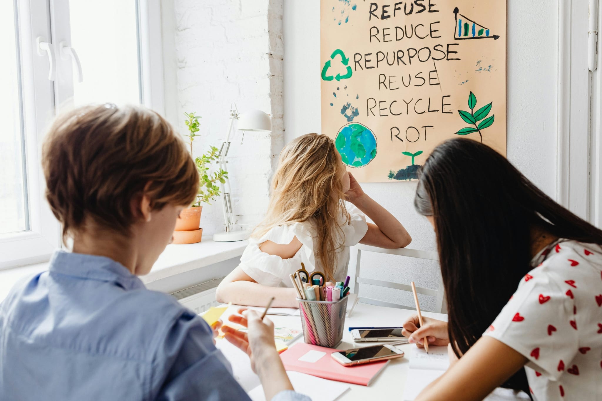 Gruppe junger Frauen sitzt an einem Tisch und arbeitet, eine junge Frau blickt nach hinten auf ein selbstgeschriebenes Plakat zu Umweltthemen ("Reuse, recycle...").