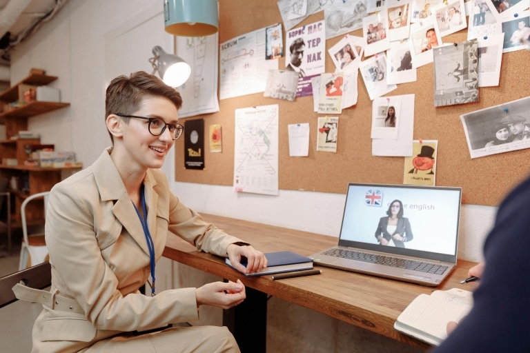Junge Frau mit kurzen Haaren und beigem Anzug sitzt am Schreibtisch, dahinter eine Pinnwand mit Fotos und Artikeln, am Tisch ein aufgeklappter Laptop Text "english"