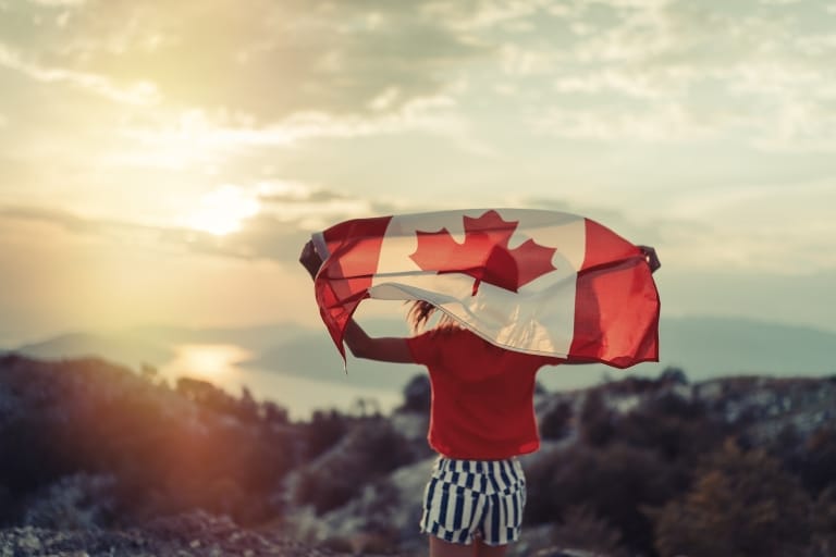 Junge Frau steht auf einem Hügel und blickt hinunter auf einen Fluss. Über dem Kopf schwenkt sie die Flagge Kanadas.