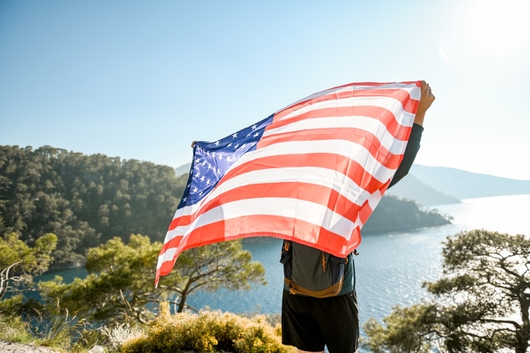 Mann, auf einer Klippe stehend, hält die amerikanische Flagge - im Hintergrund Meer und Berge.