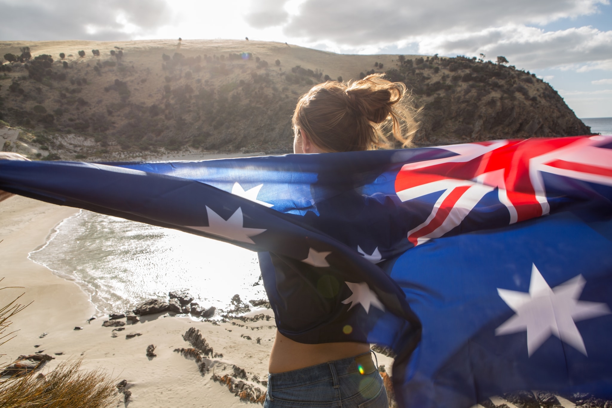 Junge Frau von hinten steht auf einer Klippe über einem Strand und hält eine große australische Flagge.