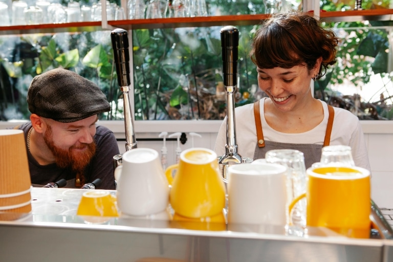 Junge Frau und junger Mann hinter einer Kaffeemaschine, beide lächeln. Im Vordergrund gelbe und weiße Kaffeehäferl.