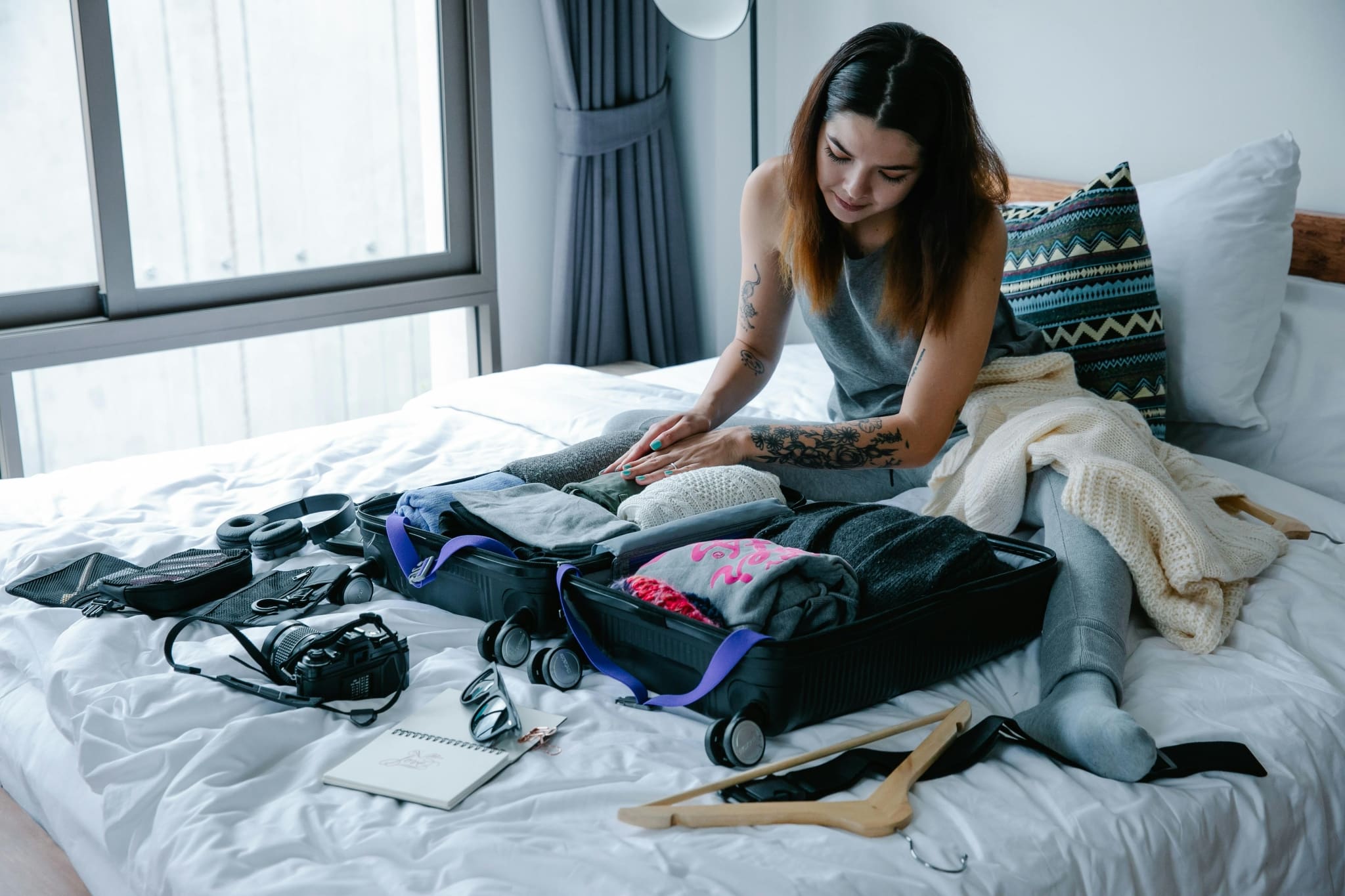 Junges Mädchen mit kurzem T-Shirt und tätowierten Unterarmen sitzt am Bett und packt ihren Koffer.