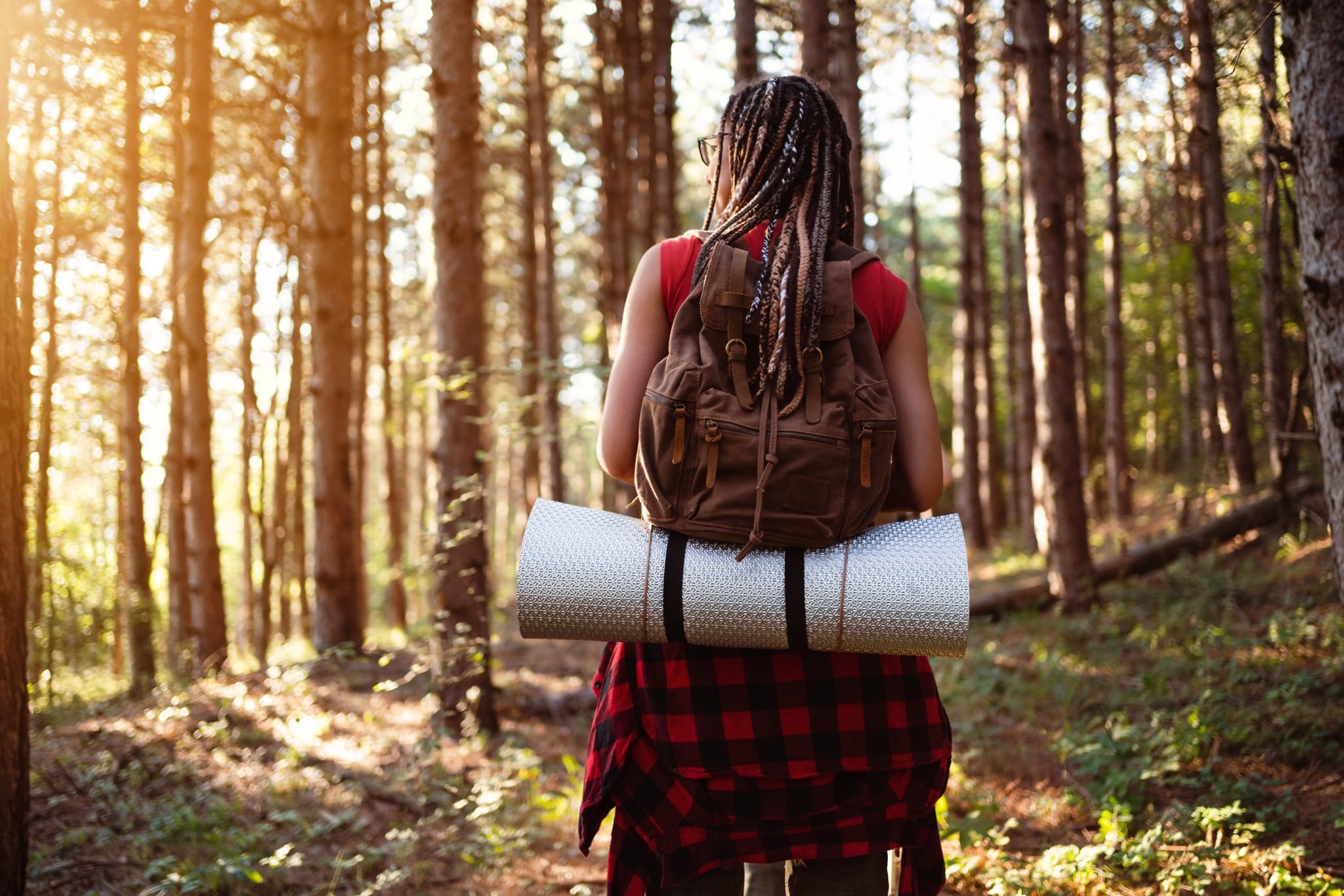 Junge Frau mit Dreads von hinten mit Rucksack und Isomatte, die durch einen Wald wandert.