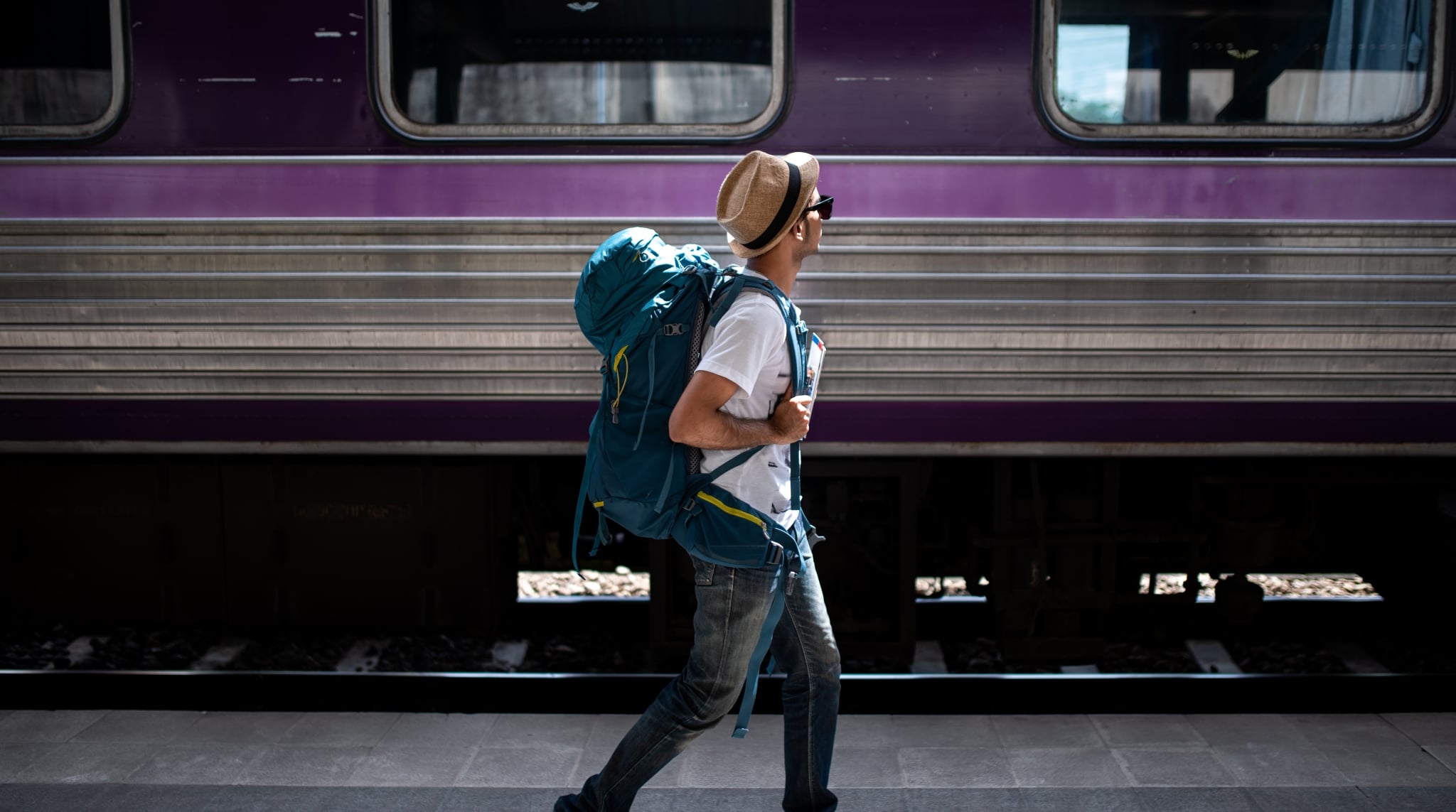 Junger Mann mit großem Tramper-Rucksack und Sonnenhut am Bahnsteig, im Hintergrund ein Zug