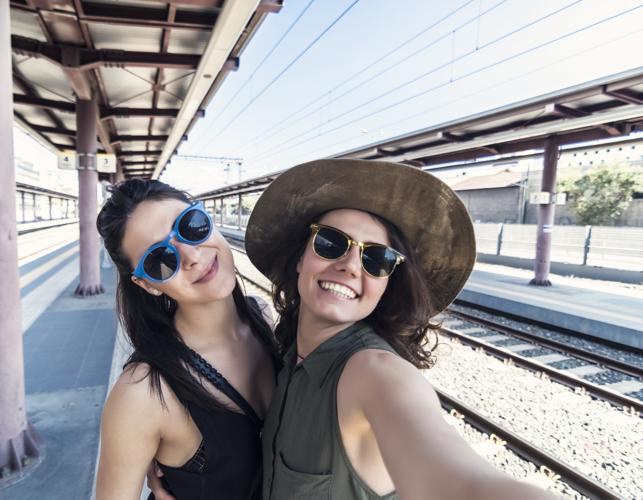 Zwei junge Frauen mit Sonnenbrillen und strahlendem Lächeln machen ein Selfie am Bahnhof, dahinter Schienen.