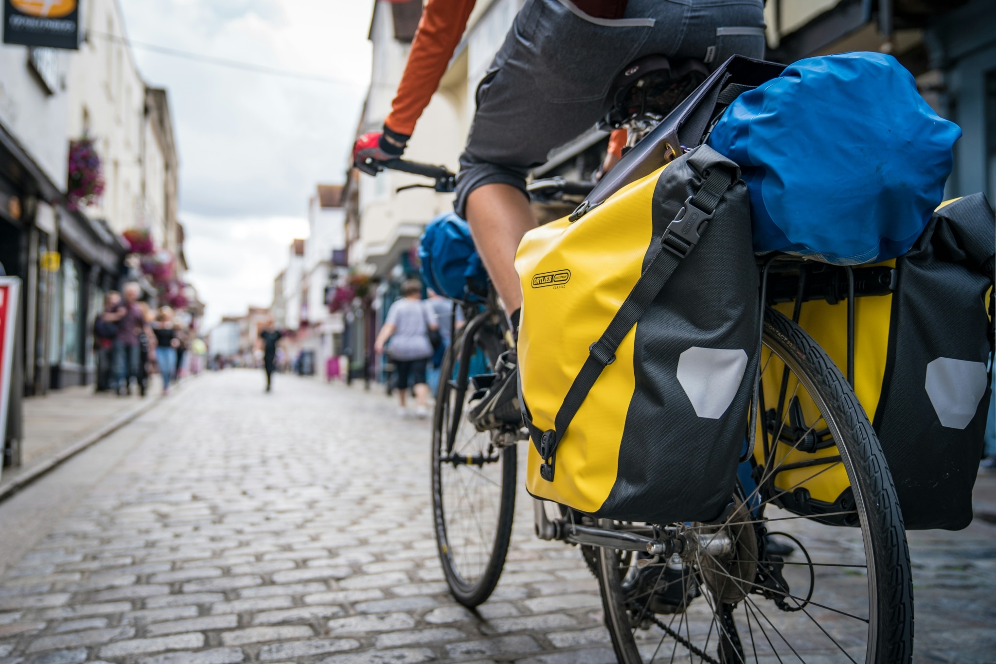Radfahrer mit gelben großen Packtaschen, der auf Straße mit Kopfsteinpflaster durch eine Stadt radelt.