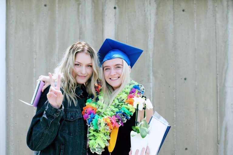 Zwei junge Mädchen beim Schulabschluss, eine davon mit blauem Hut und bunter Blumenkette um den Hals.