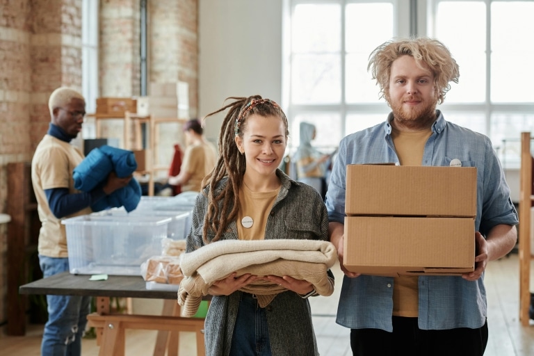 Junge Freiwillige - eine junge Frau mit Dreads und ein junger Mann mit blondem Bart - halten Decken und Kartons in der Hand.