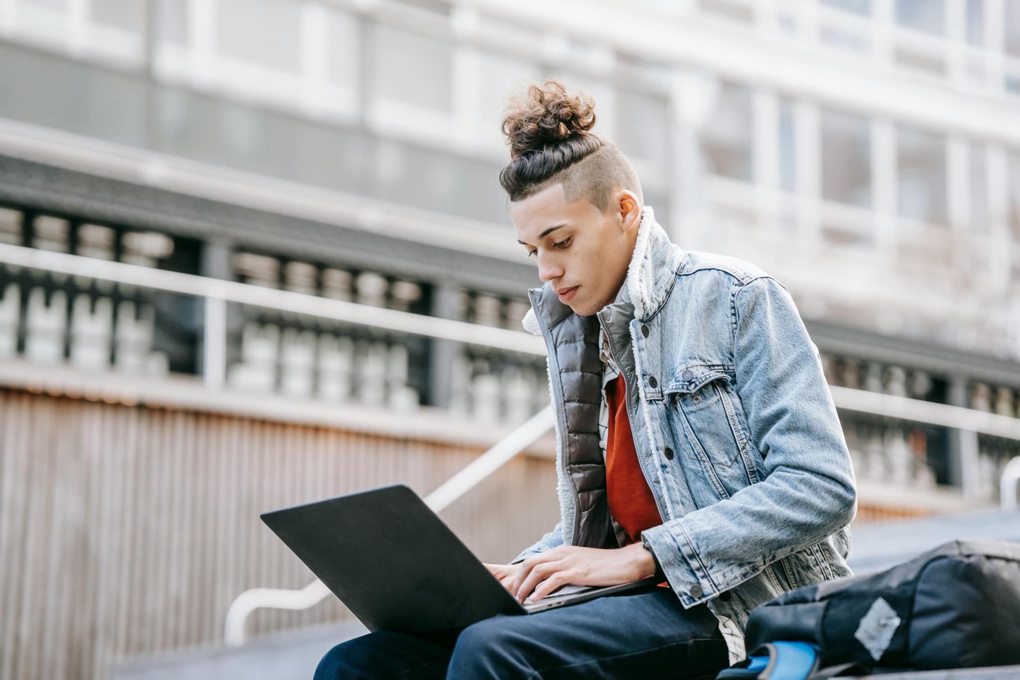 Junger Mann mit Jeansjacke sitzt im Freien auf einer Stiege und recherchiert am Laptop.