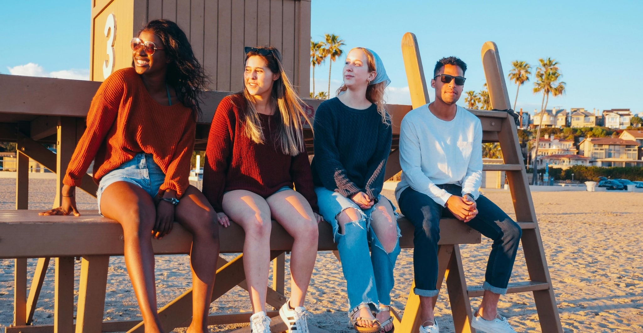 4 Jugendliche sitzen auf einem Holzgerüst am Sandstrand, lächeln und schauen in die Ferne